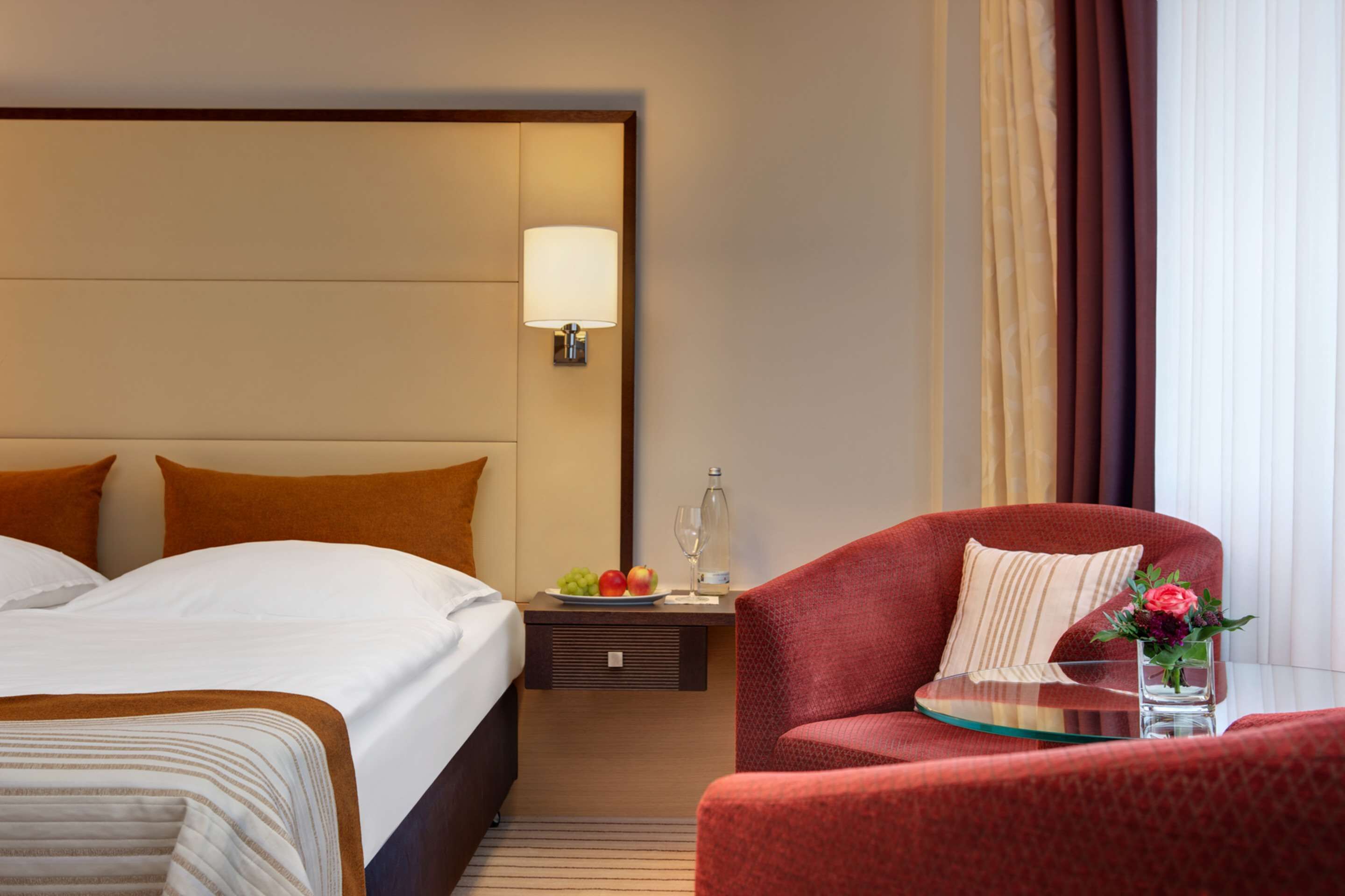 A hotel room with a large bed, a red armchair, a small table with a vase of flowers, and a nightstand with a lamp and a bottle.