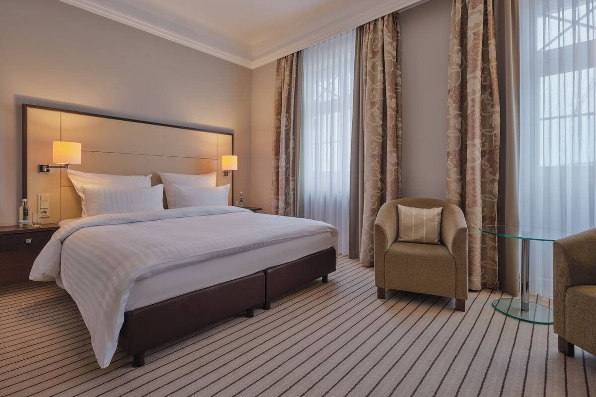 A hotel room with a large bed, two bedside lamps, a chair, and a glass table.