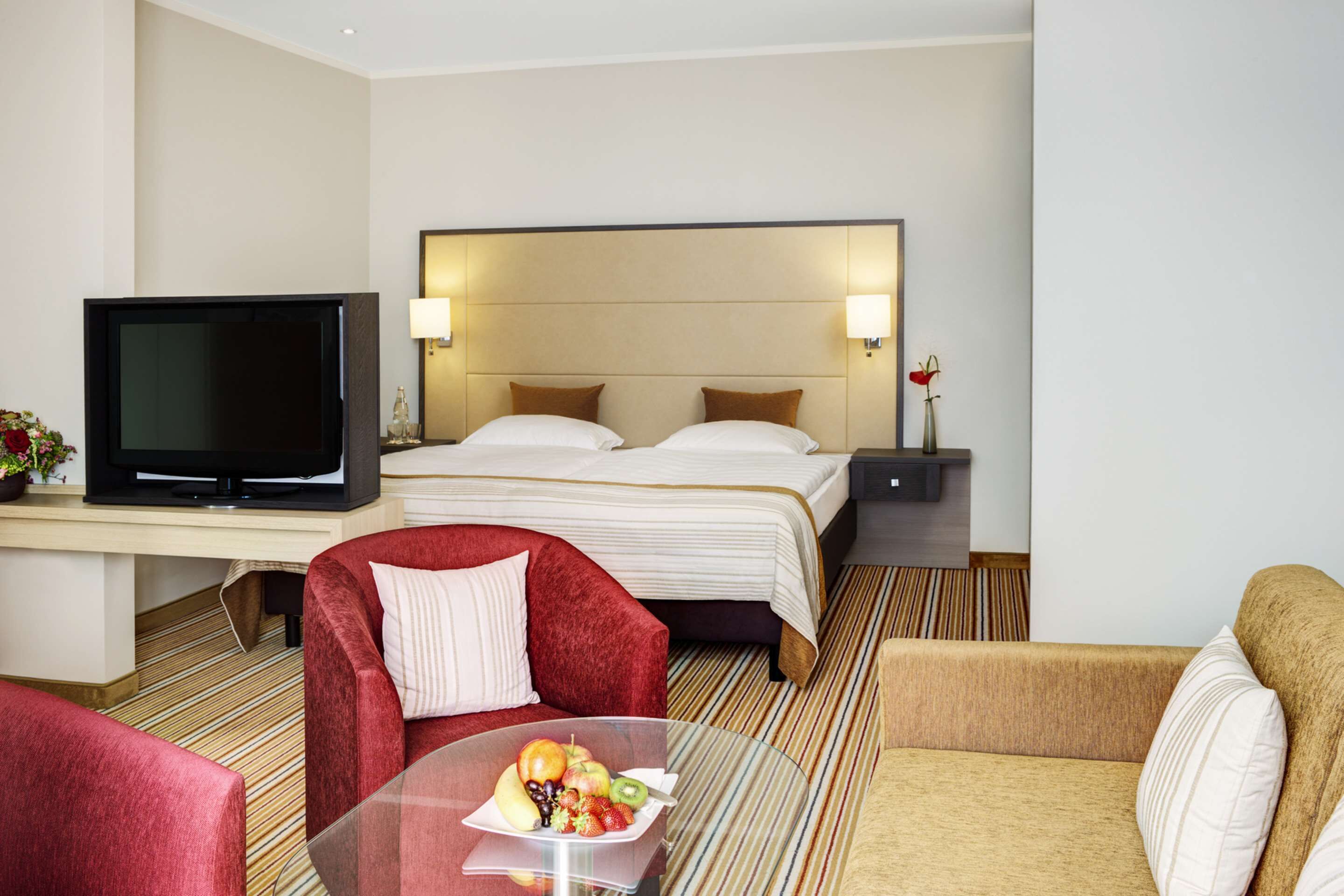 Habitación de hotel con cama, televisión, una mesita de vidrio con fruta y dos sofás.