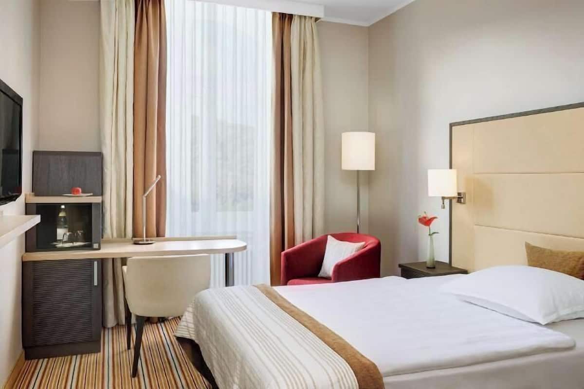A hotel room with a bed, a desk, a chair, a TV, and a lamp.