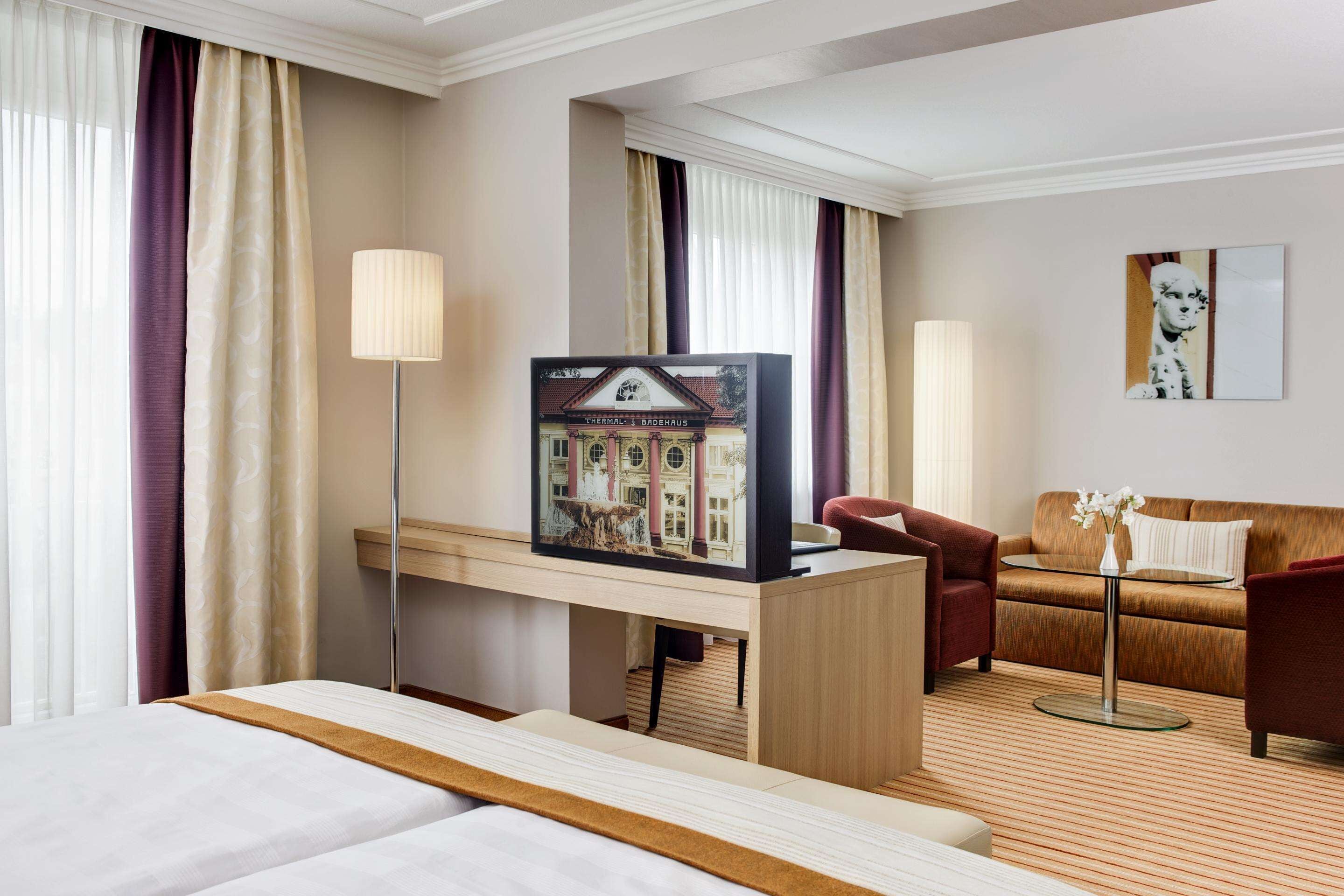 A hotel room with a bed, a desk with a lamp, a sofa, and a painting on the wall.