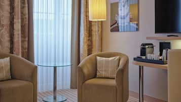 A hotel room with two armchairs, a glass table, a lamp, a coffee maker, and a TV on a stand.