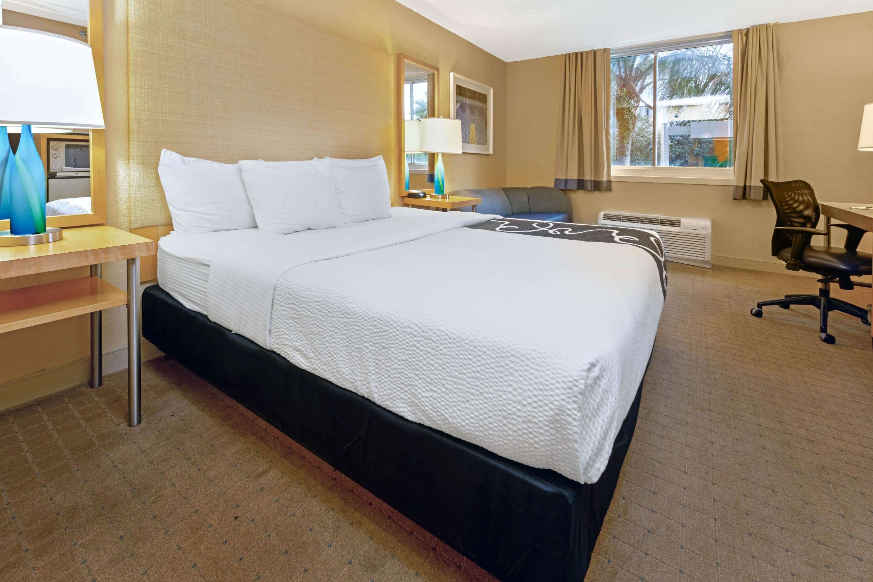 A hotel room with a large bed, a desk with a lamp, a chair, and a window with curtains.