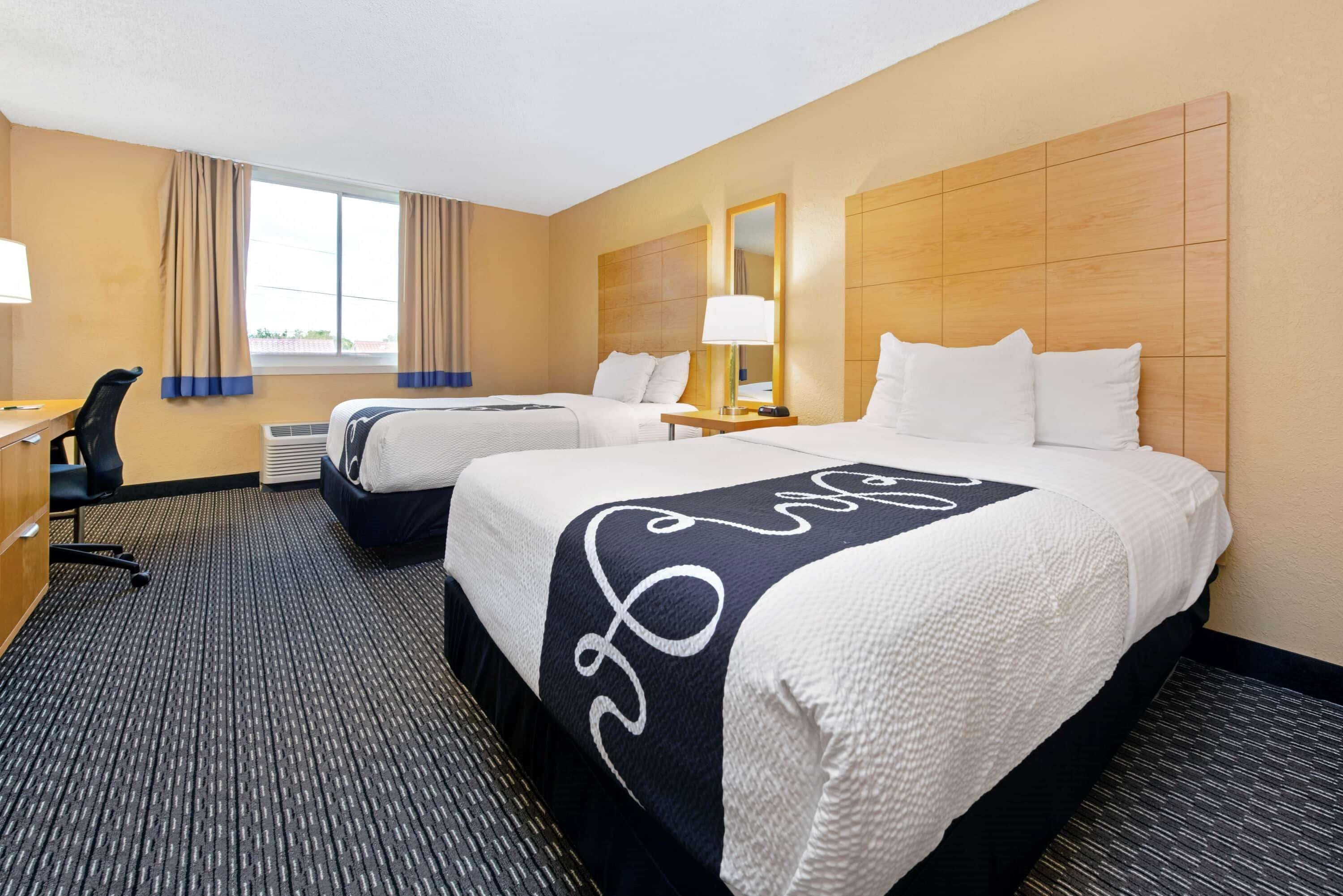 A hotel room with two beds, a desk, a chair, and a large window with curtains.