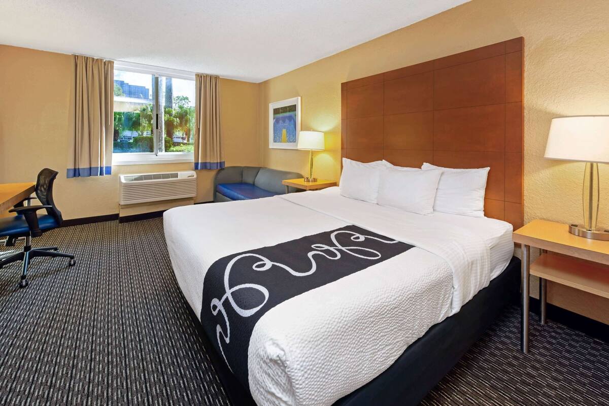 A hotel room with a large bed, a desk, a chair, a window with curtains, and a wall-mounted lamp.