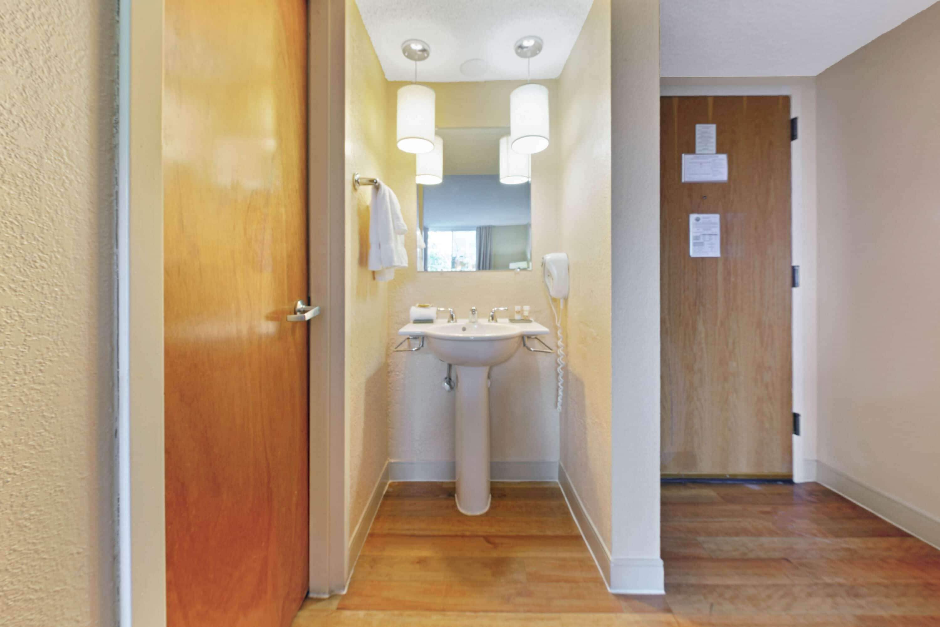 Un pasillo estrecho con una puerta de madera a la izquierda, un lavamanos blanco con un espejo encima y otra puerta de madera al final.
