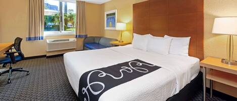 A hotel room with a large bed, a desk, a chair, a window with curtains, and a wall-mounted lamp.