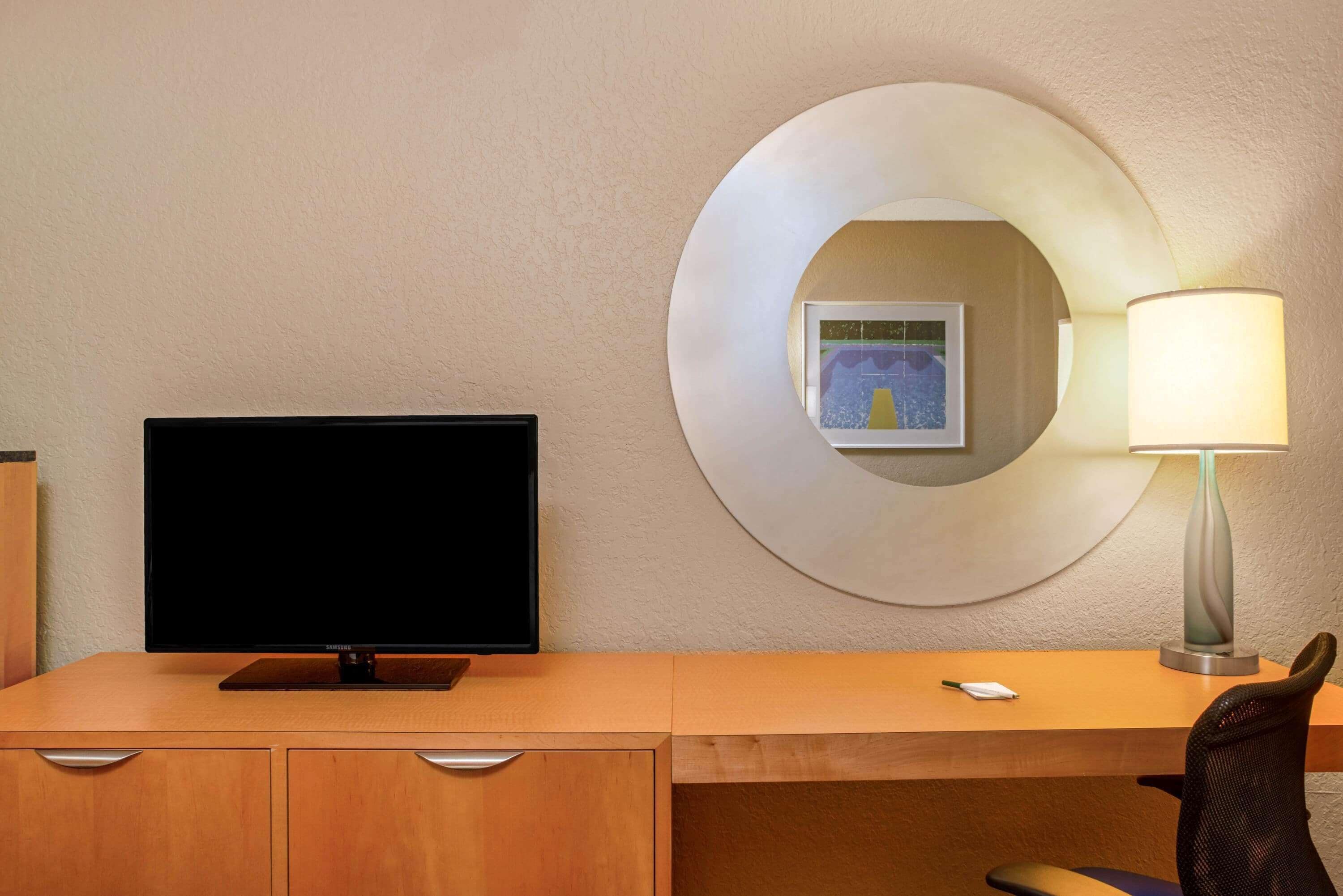 Une chambre d’hôtel avec un bureau en bois, un téléviseur à écran plat, une lampe moderne et un tableau encadré au mur.