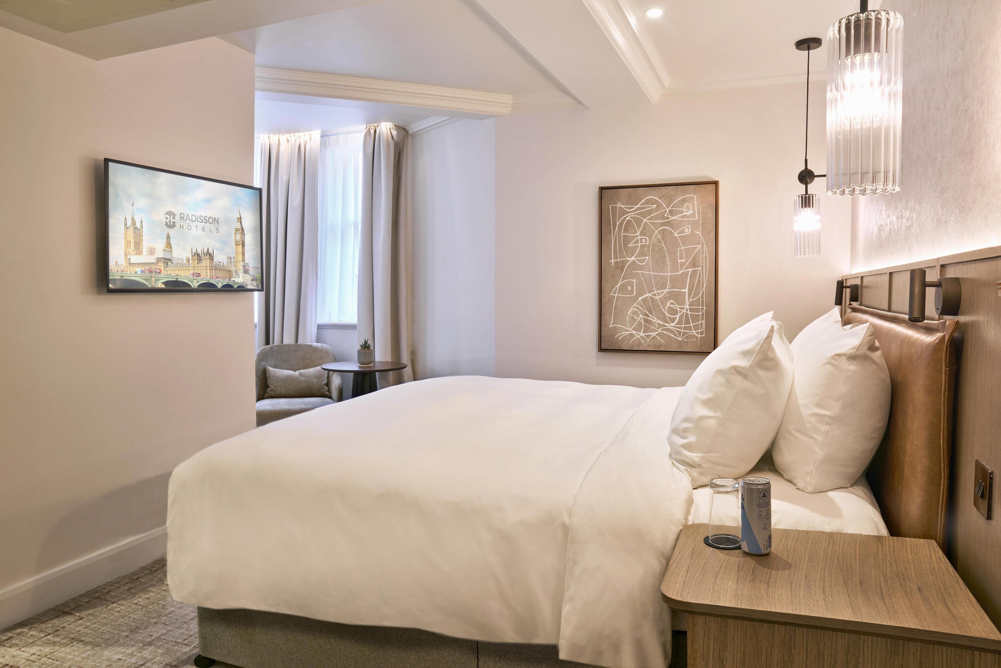 Et hotelværelse med en seng, et sengebord med en dåse, en lampe, et billede af en bygning og et vindue med gardiner.
