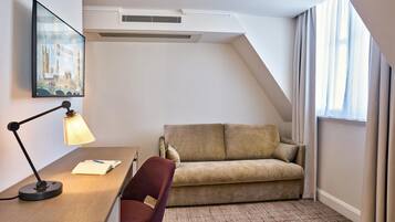 A hotel room with a desk, a chair, a sofa, a framed picture of a cityscape, and a window with curtains.