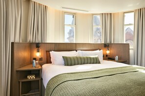 Een hotelkamer met een groot bed, twee nachtkastjes, een groene plaid en uitzicht op een gebouw door de ramen.
