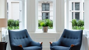Una habitación con dos sillones azules, una planta en maceta y vistas a un edificio de ladrillo a través de amplias ventanas.