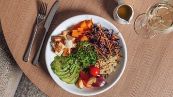 A plate of food with various vegetables and grains, a fork and knife, a cup of coffee, and a glass of white wine on a wooden table.