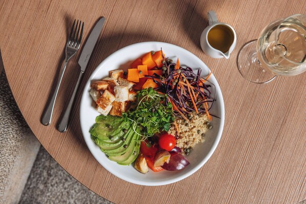 A plate of food with various vegetables and grains, a fork and knife, a cup of coffee, and a glass of white wine on a wooden table.