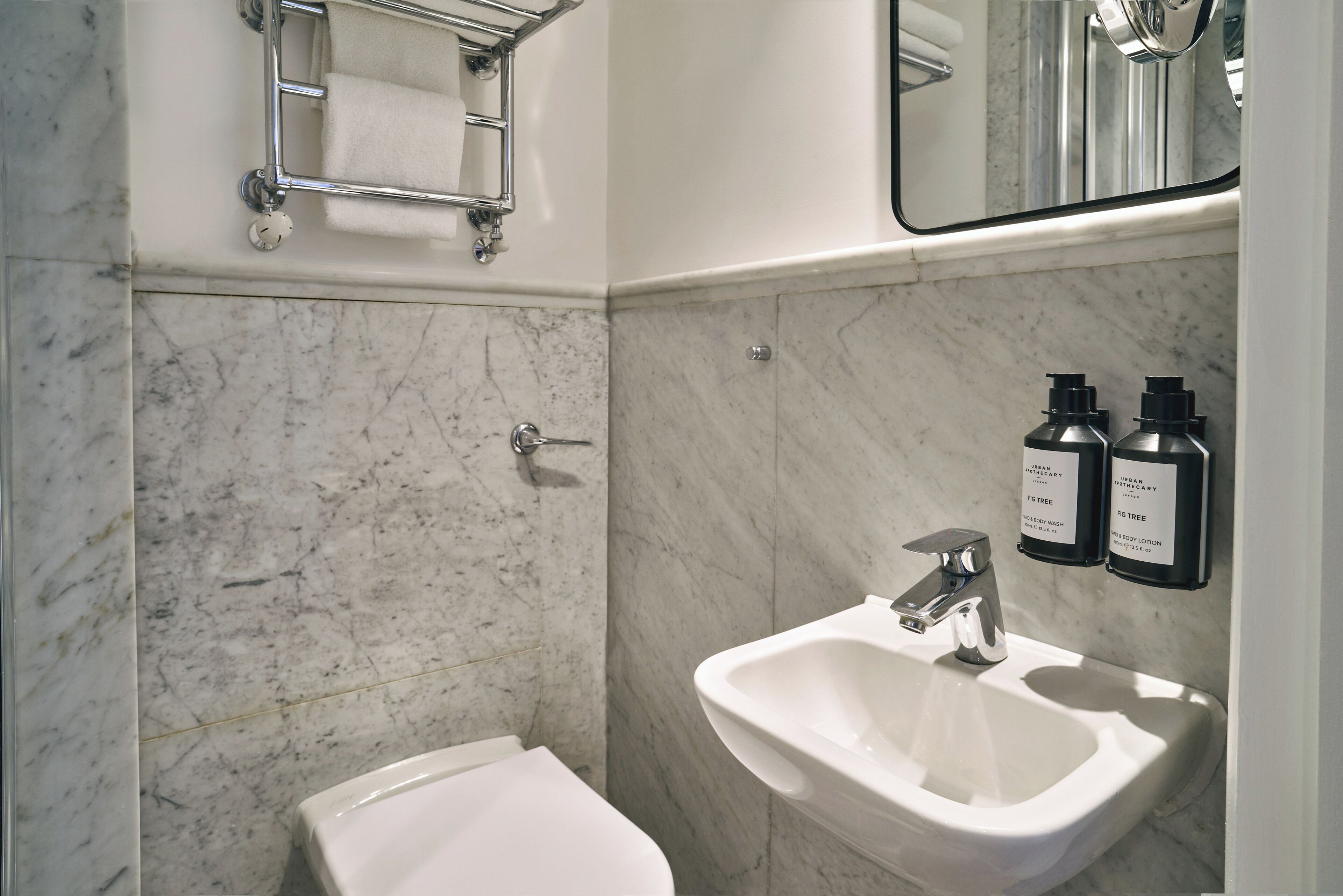 Un baño con inodoro blanco, lavamanos y espejo. Hay dos botellas de jabón corporal y loción en la pared.