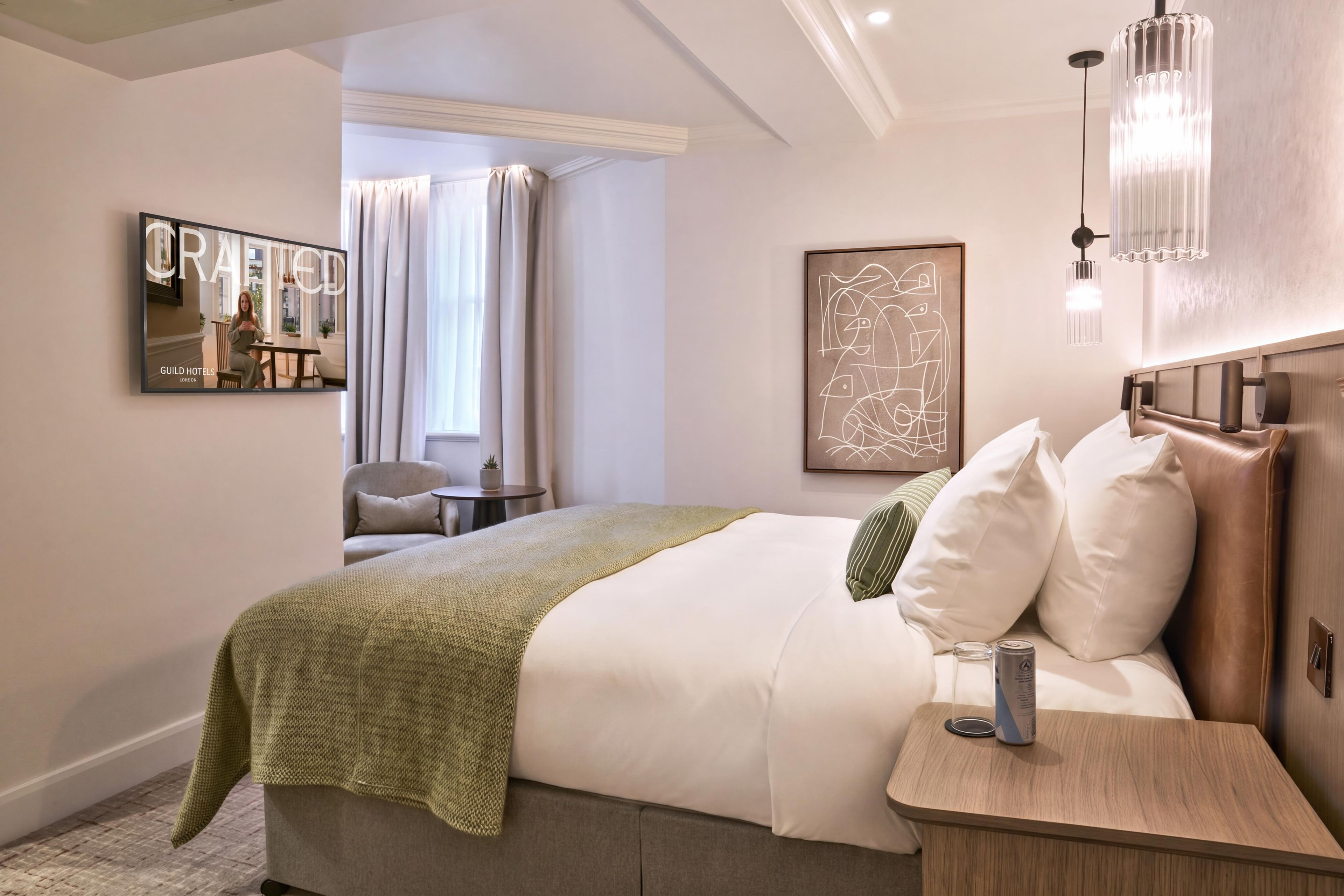 A hotel room with a bed, bedside table, lamp, artwork, and a view of the outside.