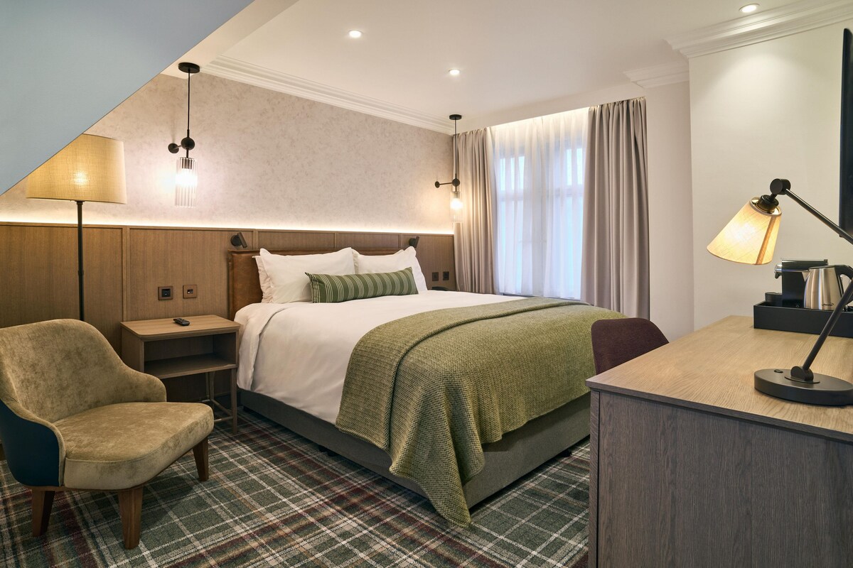 Une chambre d’hôtel avec un lit, un bureau, une chaise, une lampe et une fenêtre avec des rideaux.