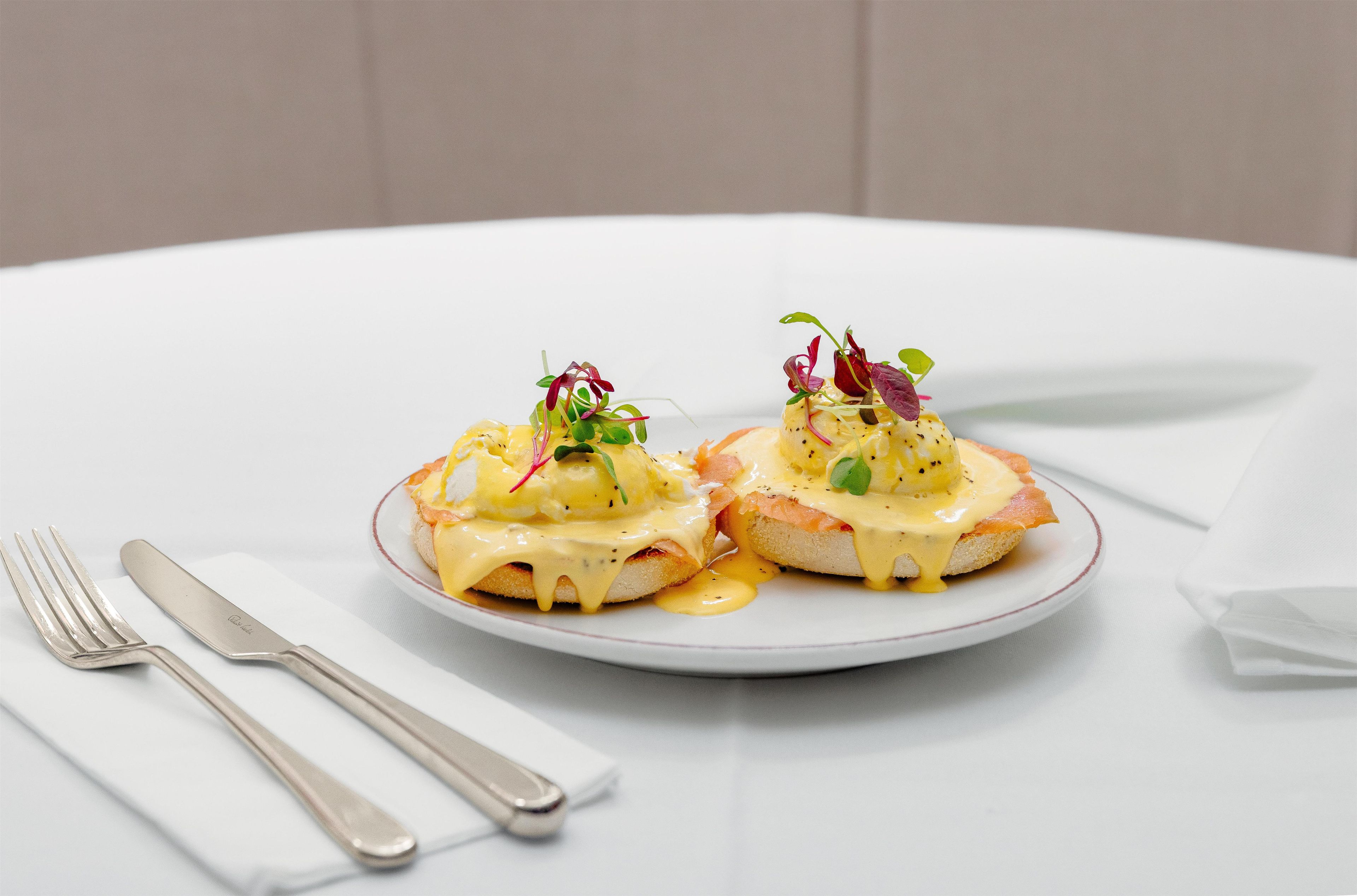 A plate of eggs benedict with hollandaise sauce and garnishes, accompanied by a fork and knife on a white table.