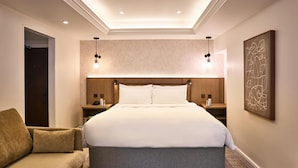 Habitación de hotel con una cama grande, dos mesitas de noche, un sofá, una obra de arte en la pared e iluminación empotrada en el techo.