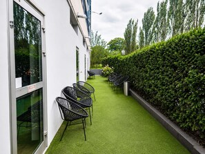 Een pad met kunstgras, zwarte metalen stoelen en een haag.