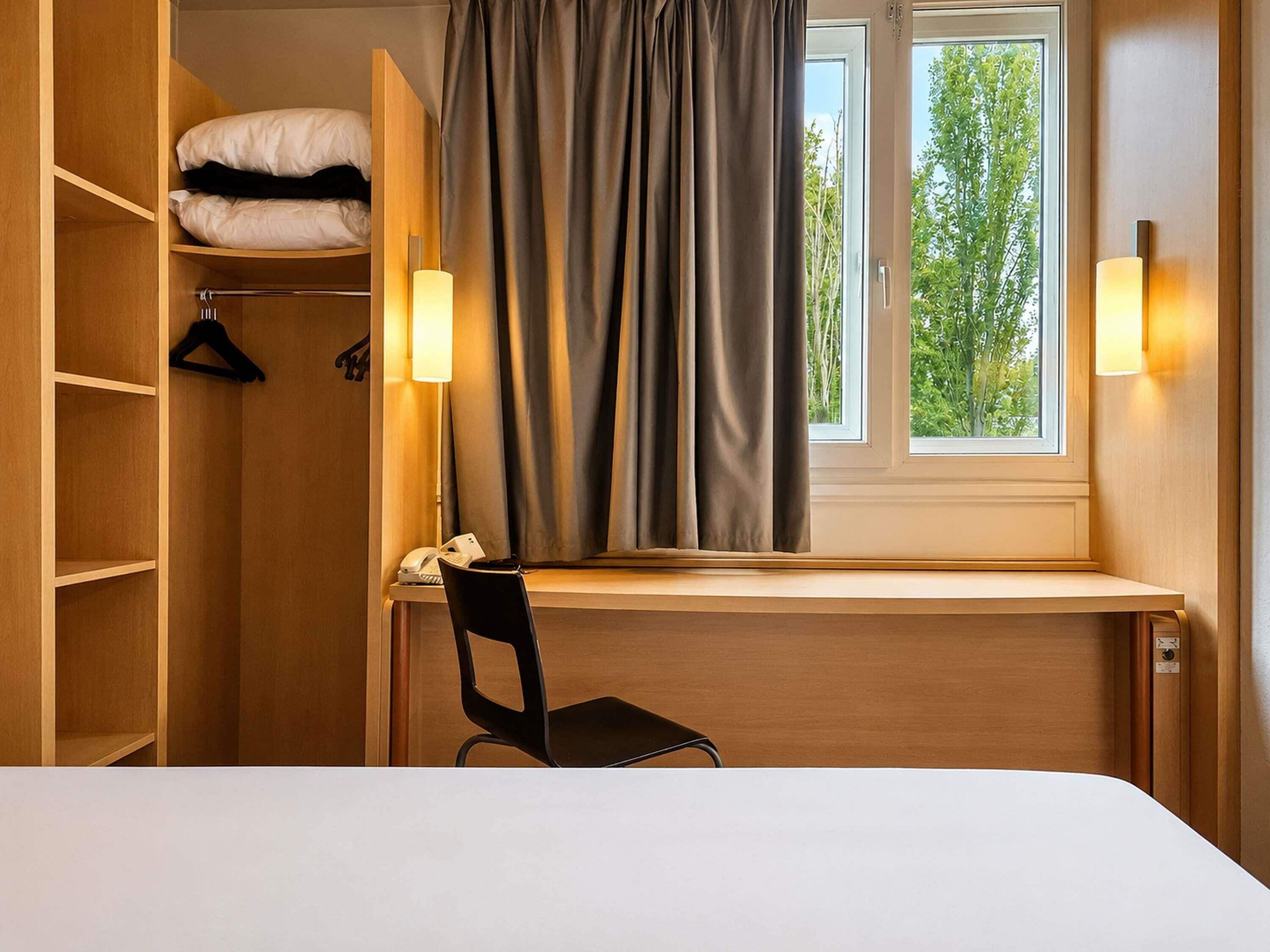Habitación de hotel con escritorio de madera, una silla, cama, armario y ventana con cortinas.