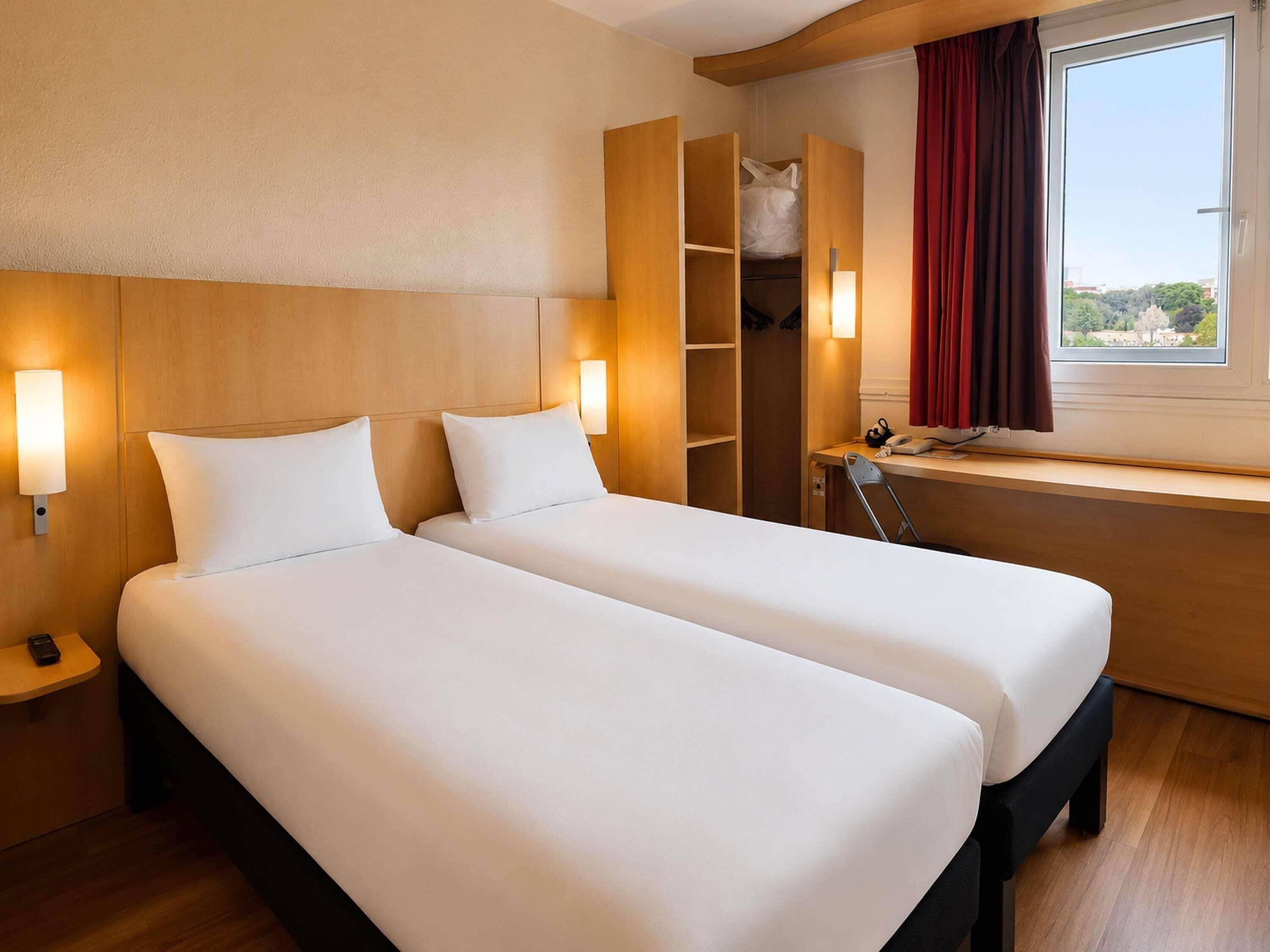 A hotel room with two beds, a desk, a chair, a wardrobe, and a window with a view.