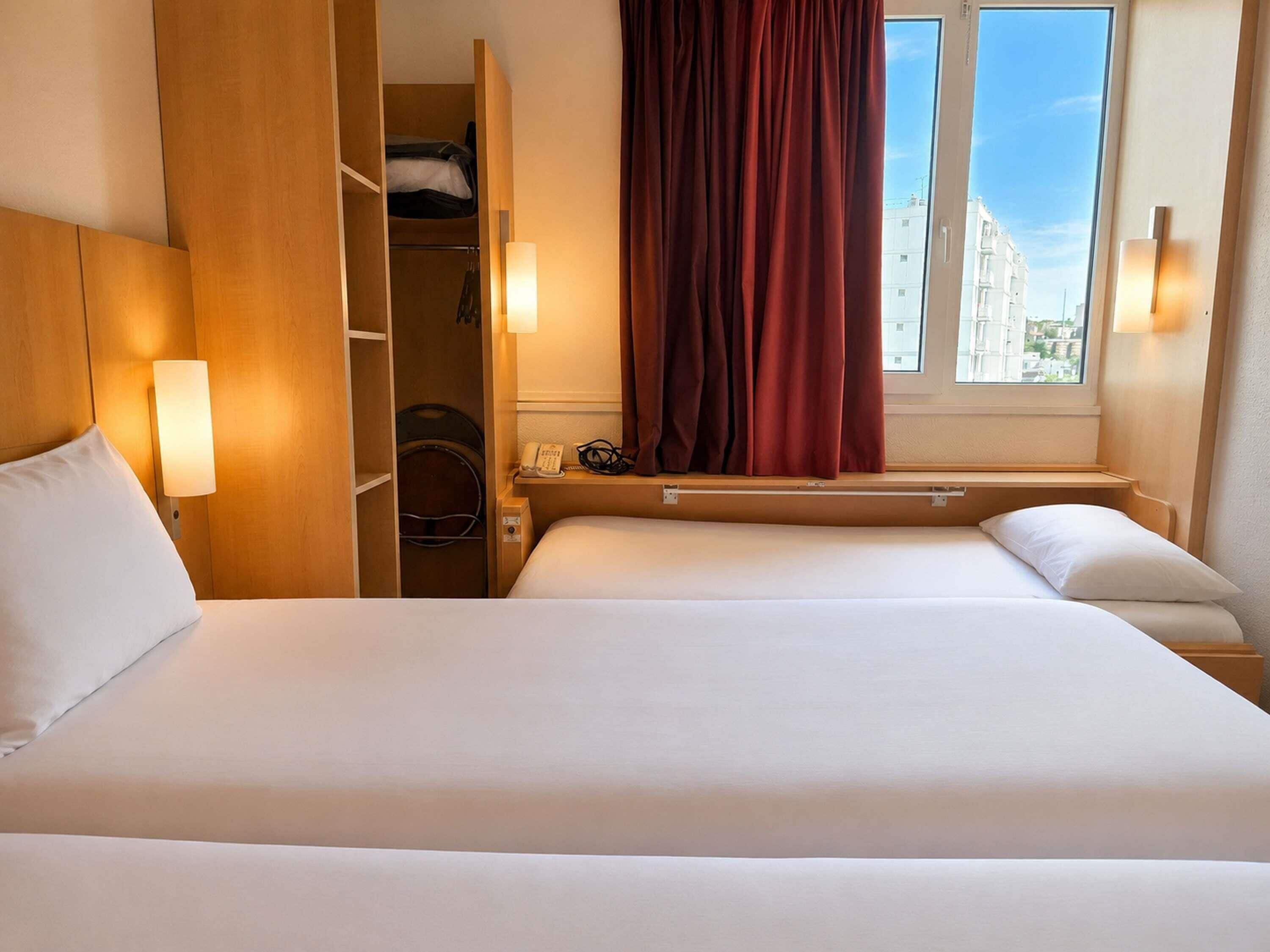 Una habitación de hotel con cama, armario, escritorio, teléfono y ventana con cortinas.