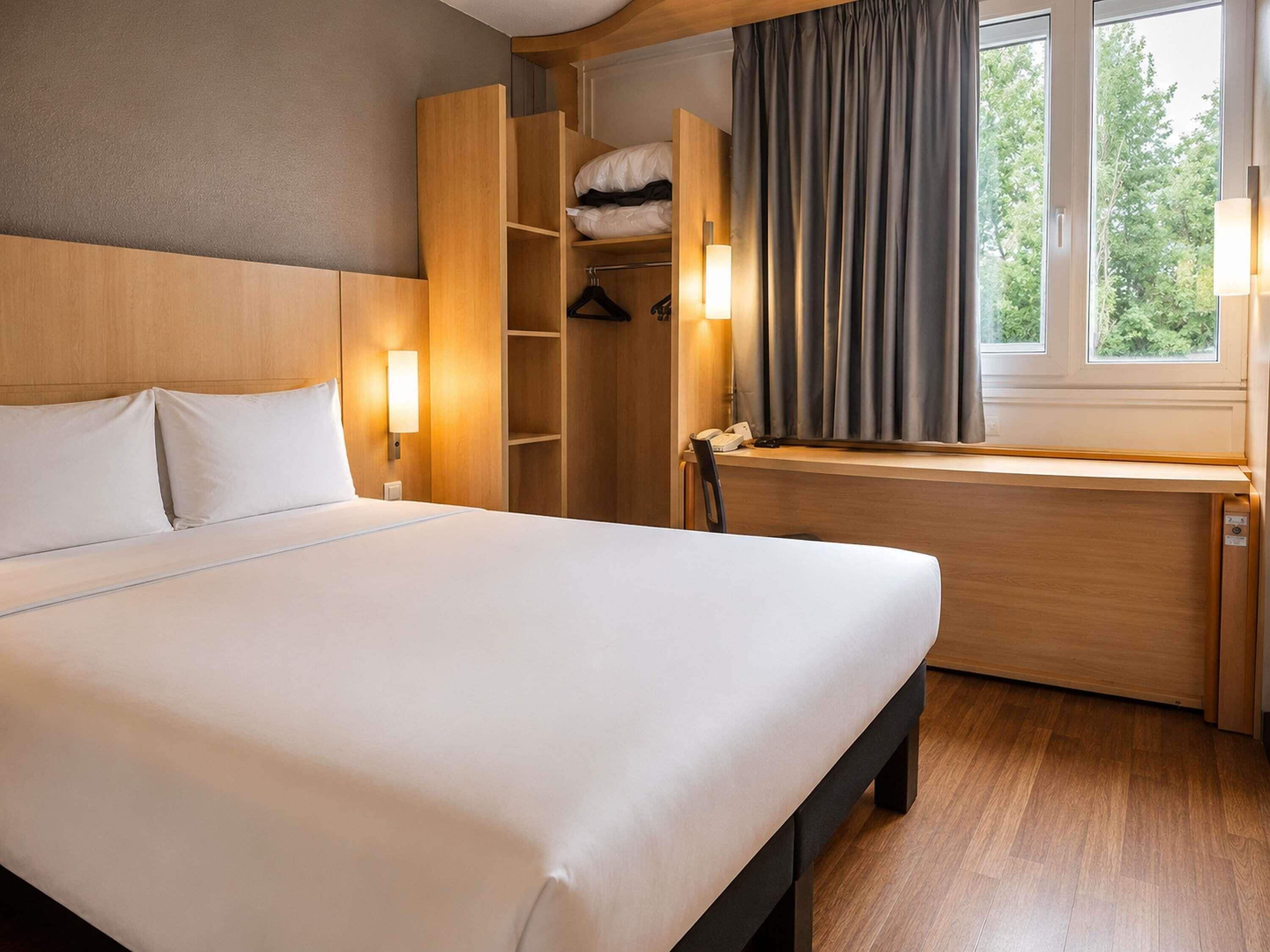 Una habitación de hotel con cama, armario de madera, escritorio y ventana con cortinas.