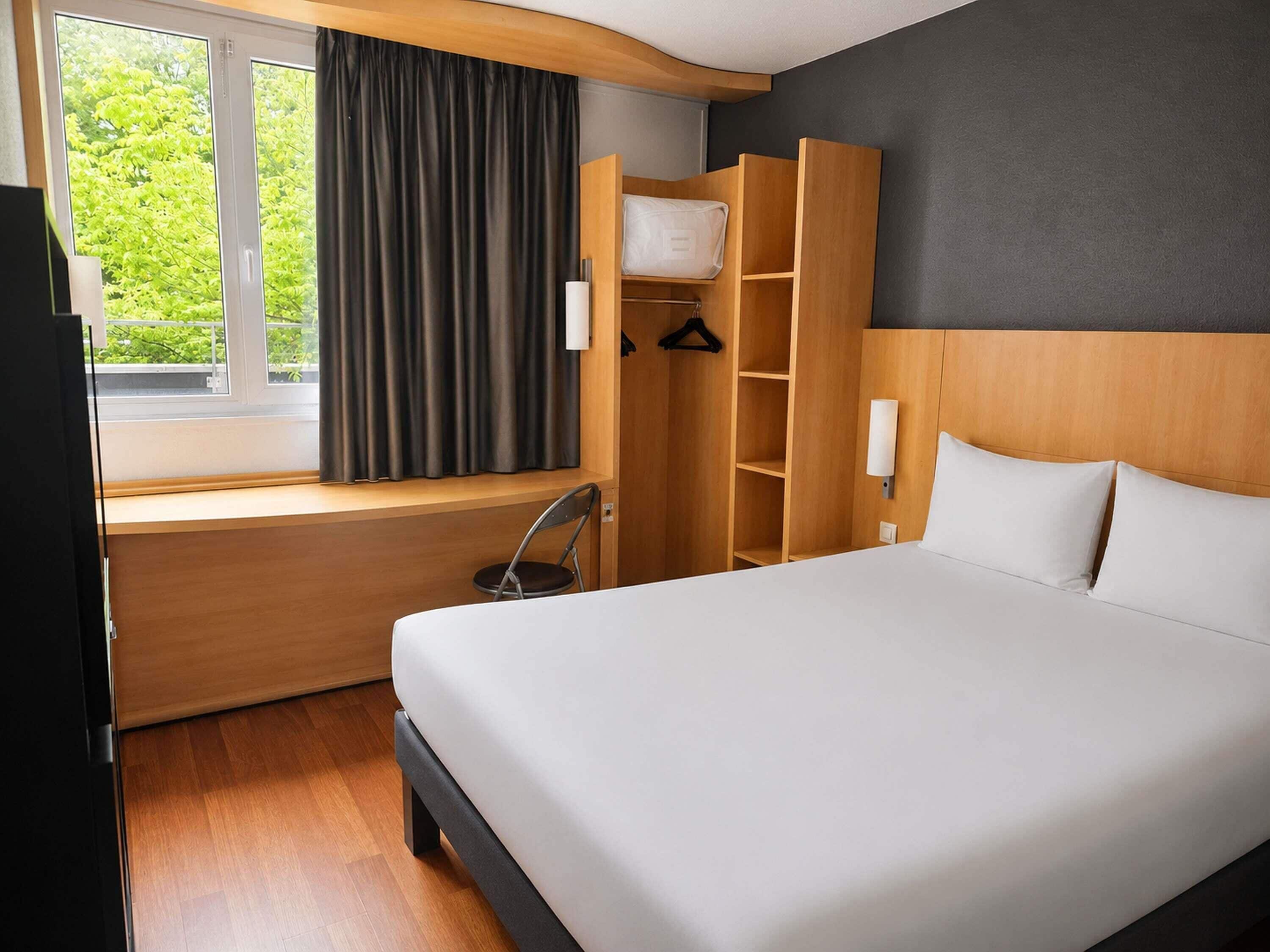 Una habitación de hotel con cama, ventana con cortinas, armario de madera y una silla.
