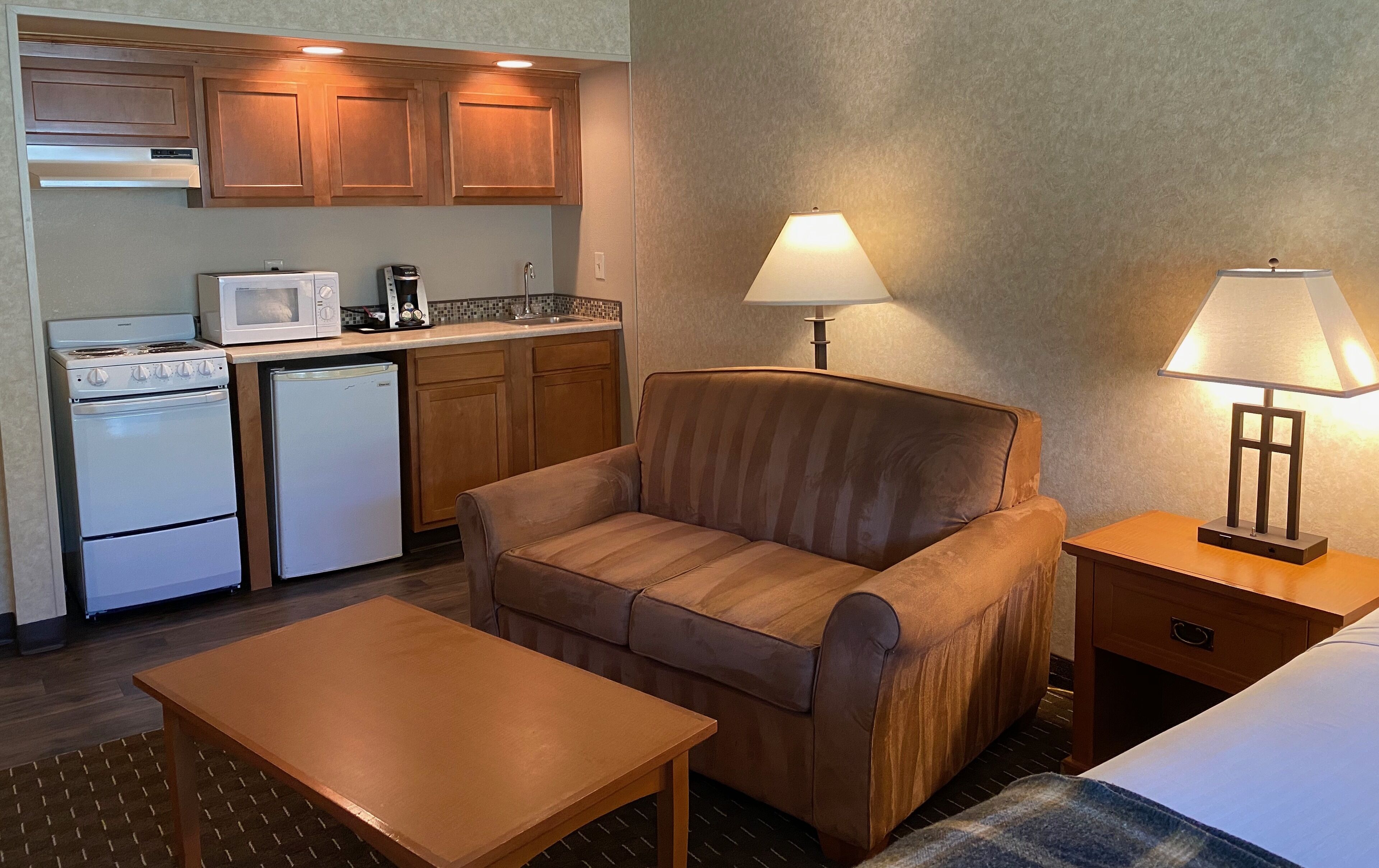 A compact hotel room with a kitchenette, a sofa, a table, and two bedside lamps.