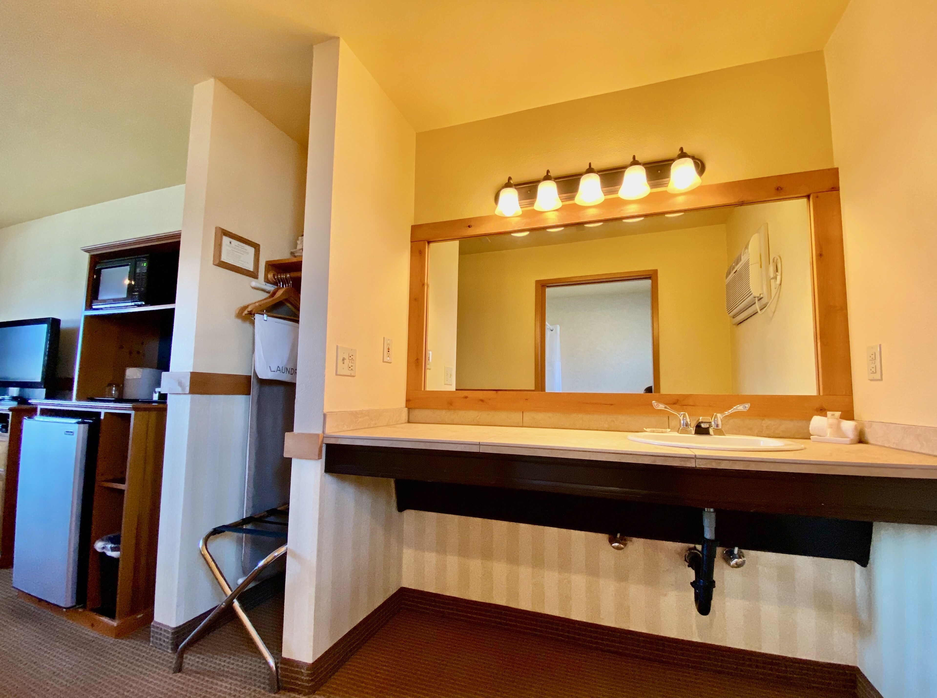 A hotel bathroom with a large mirror, a sink, and a vanity. There is a microwave and a refrigerator in the adjacent room.