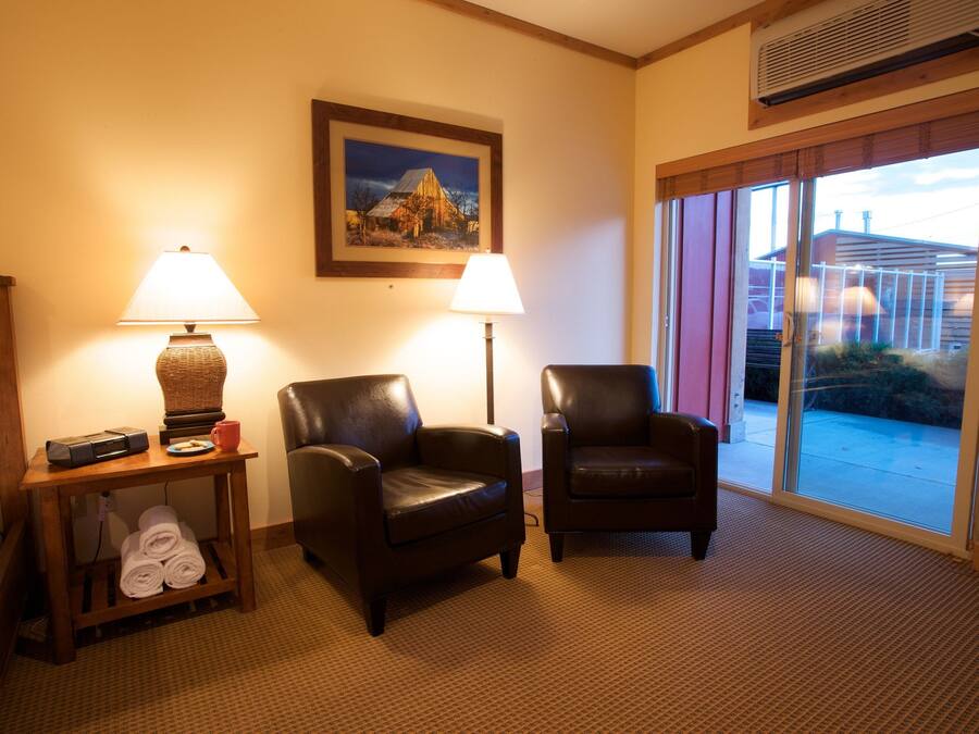 A hotel room with two leather armchairs, a small table with a lamp, a framed picture on the wall, and a view through a sliding glass door.
