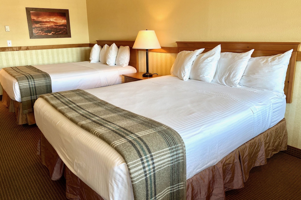 A hotel room with two beds, each with white linens and a plaid blanket. There is a nightstand with a lamp between the beds.