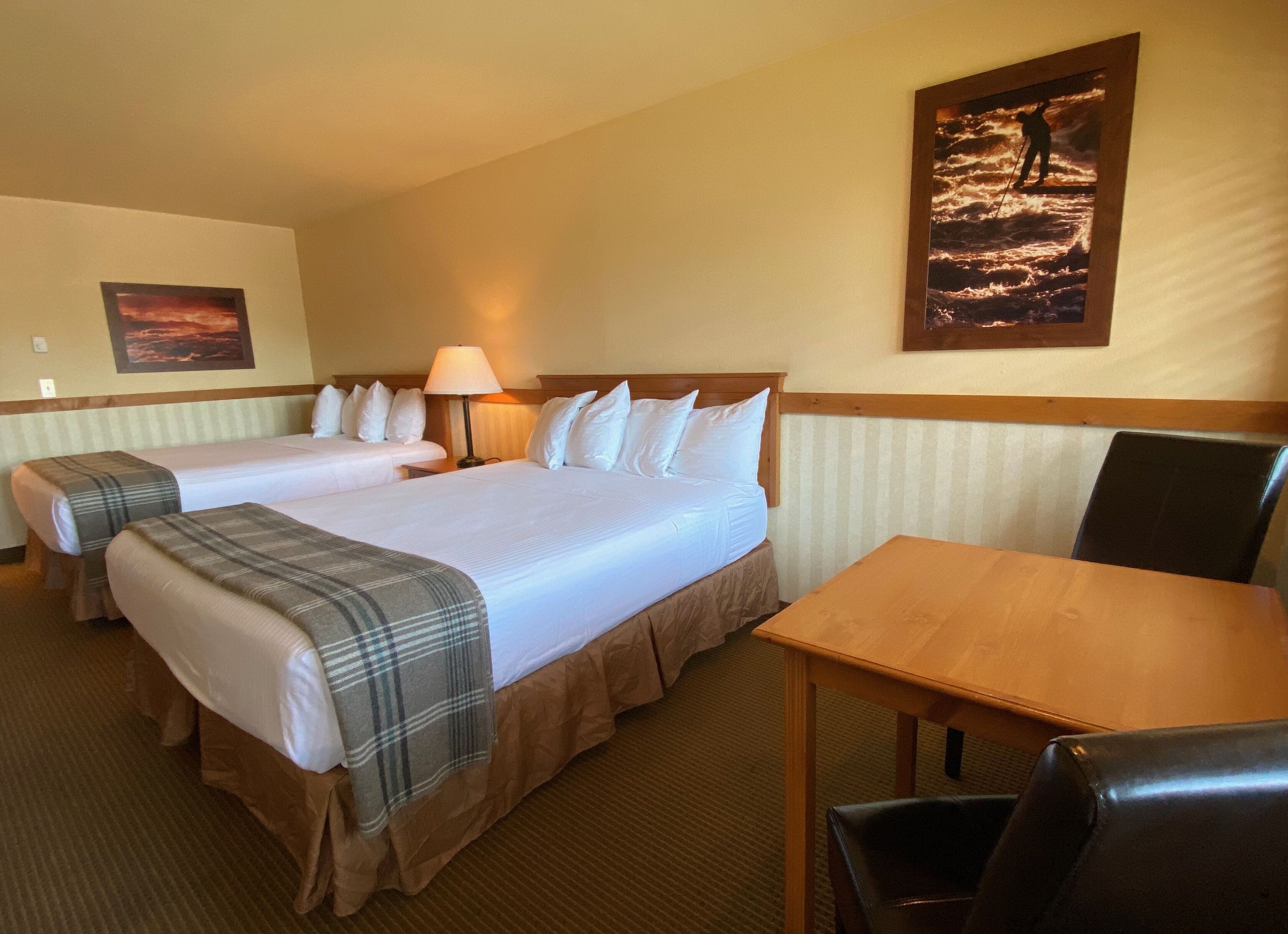 A hotel room with two beds, a wooden table, a chair, a lamp, and a painting on the wall.