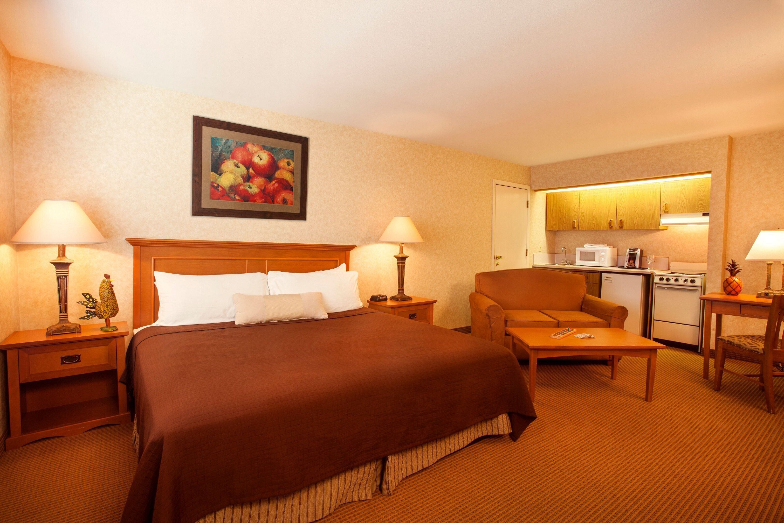 A hotel room with a large bed, a sofa, a small table, a lamp, a painting, and a kitchenette.