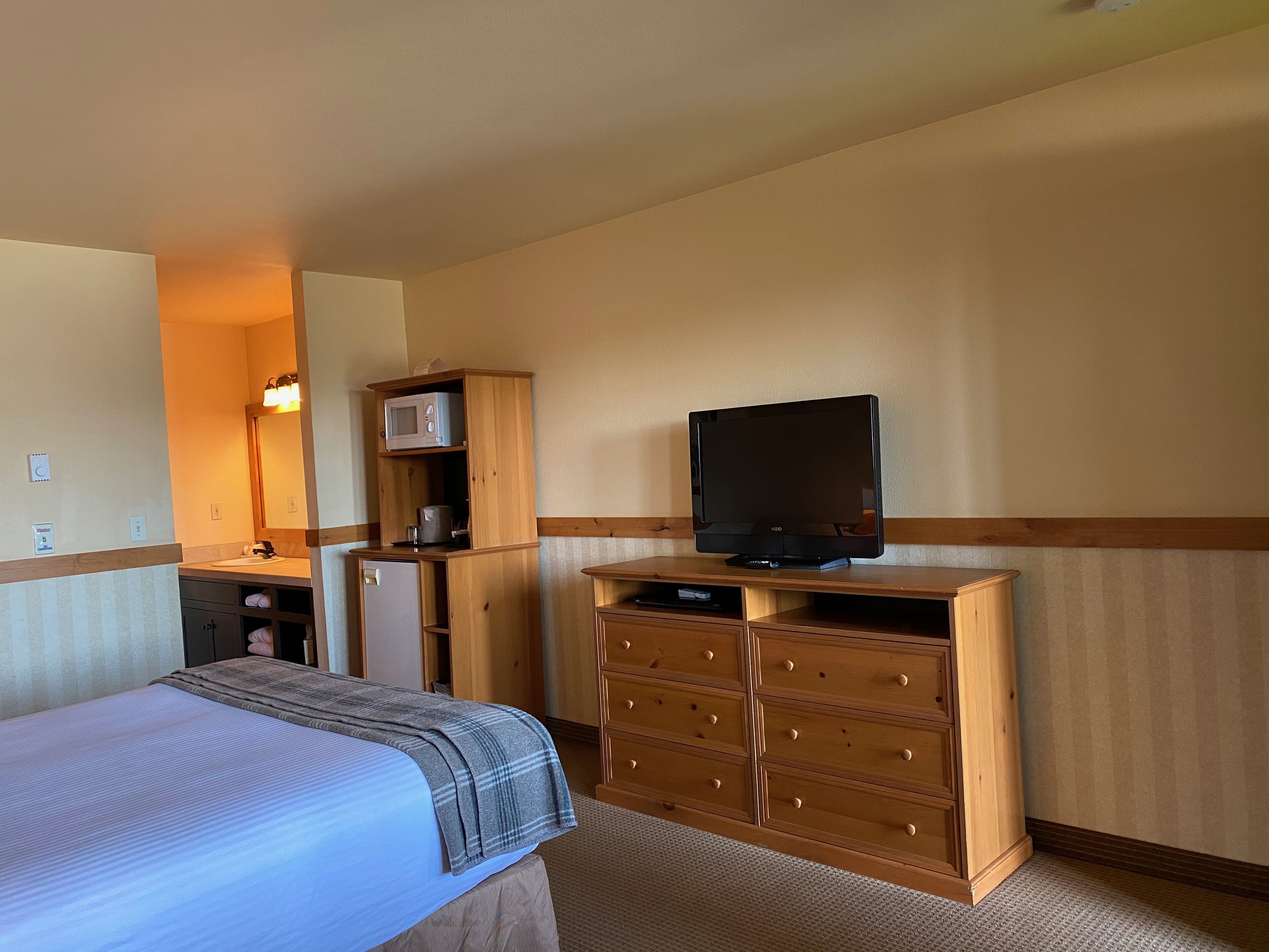 Habitación de hotel con cama, tocador de madera, televisor, microondas y una pequeña kitchenette.