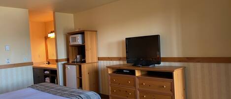 Habitación de hotel con cama, tocador de madera, televisor, microondas y una pequeña kitchenette.