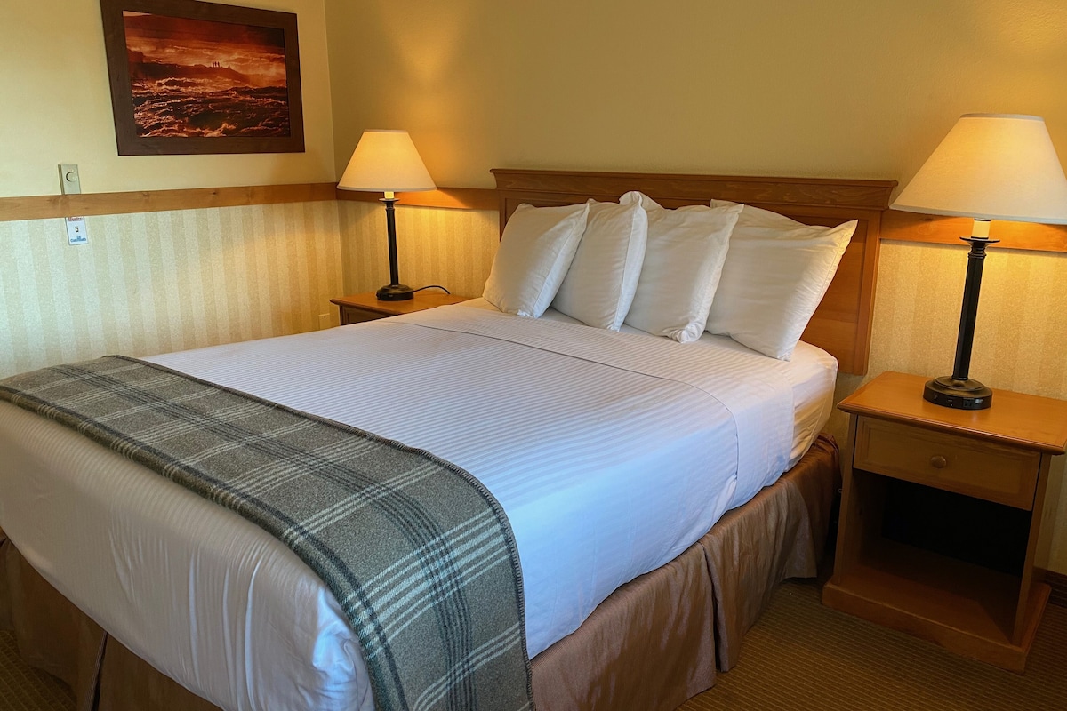 A neatly made bed with white linens and a plaid throw blanket, flanked by two bedside tables with lamps, in a hotel room with a wooden headboard and a framed picture on the wall.