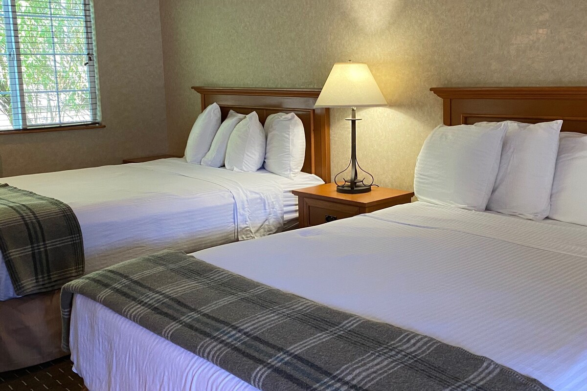 A hotel room with two beds, each with white linens and a plaid blanket. There is a nightstand with a lamp between the beds.