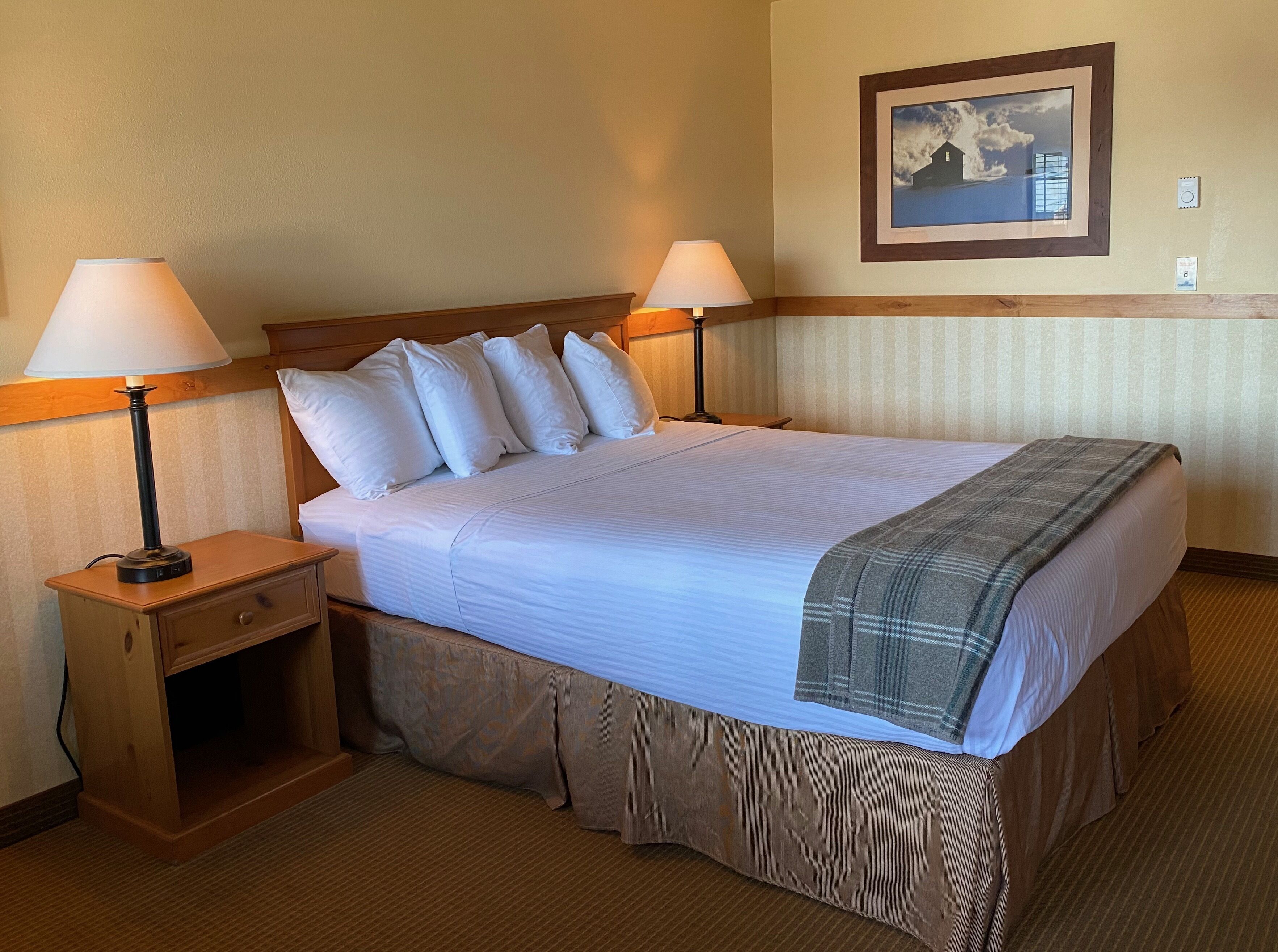 A hotel room with a large bed, two bedside lamps, a nightstand, a wooden headboard, a framed picture on the wall, and a carpeted floor.