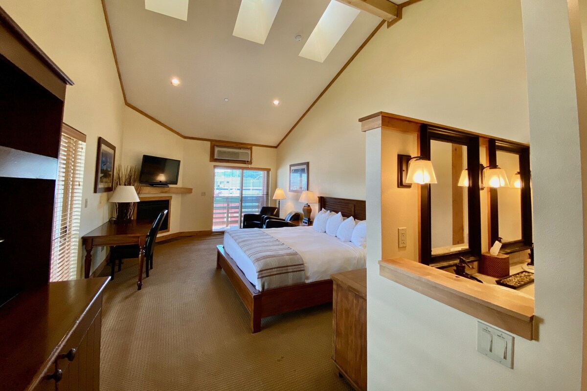 A spacious bedroom with a large bed, a desk, a television, and a bathroom with a mirror and lights.