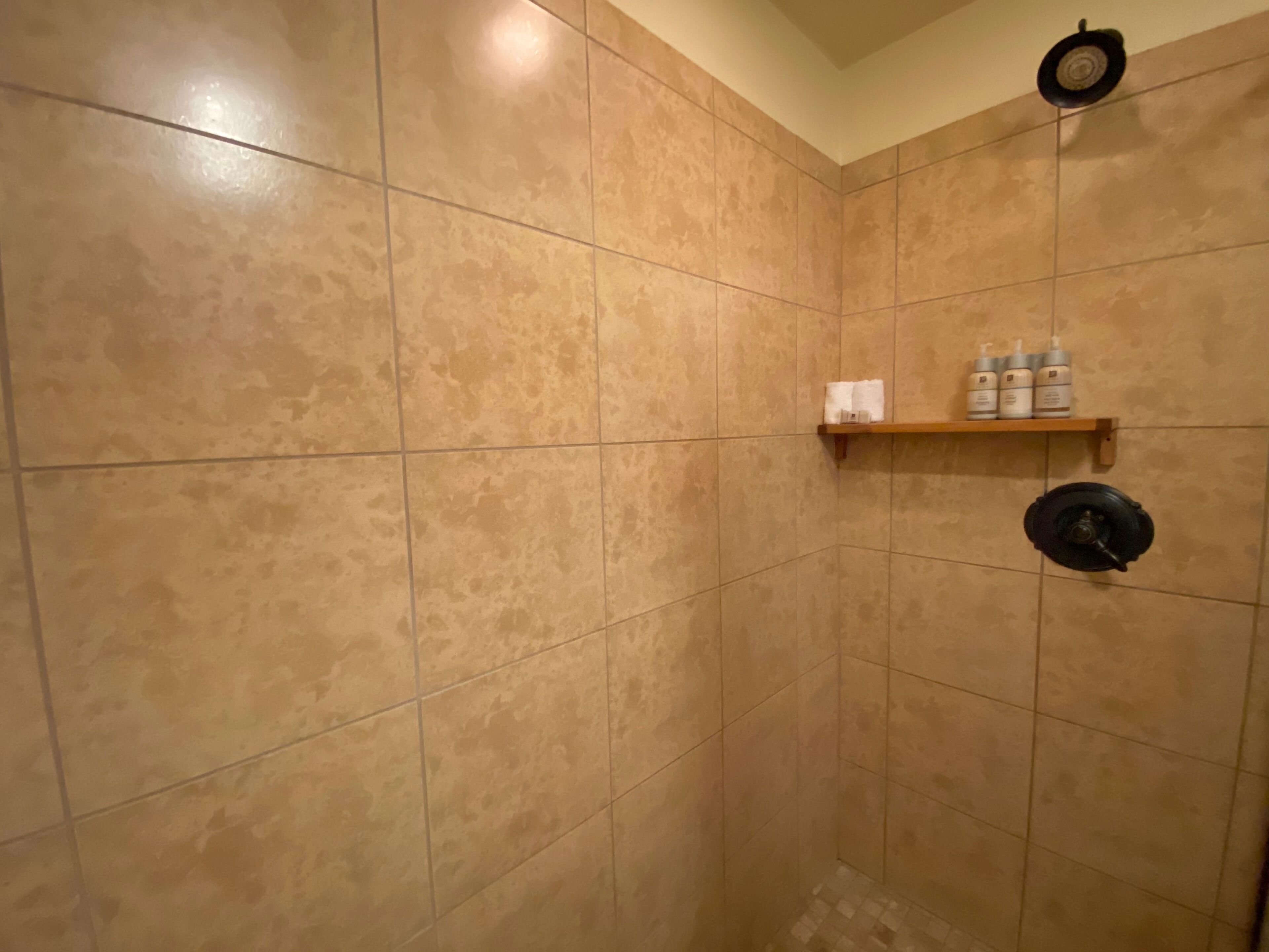 A shower area with beige tiles, a shelf with toiletries, and a showerhead.