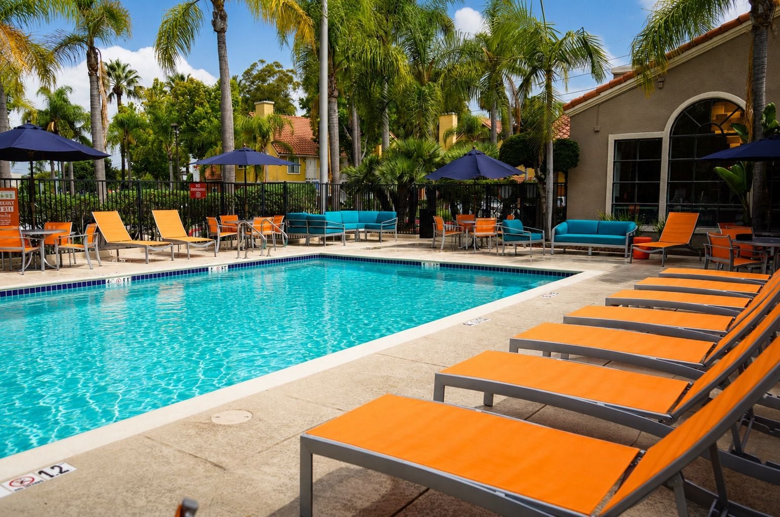 A swimming pool with lounge chairs and umbrellas, surrounded by palm trees and a building with large windows.