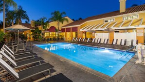 A swimming pool with lounge chairs and umbrellas, surrounded by a building with a tiled roof and palm trees. - Clementine Hotel & Suites Anaheim (Anaheim)