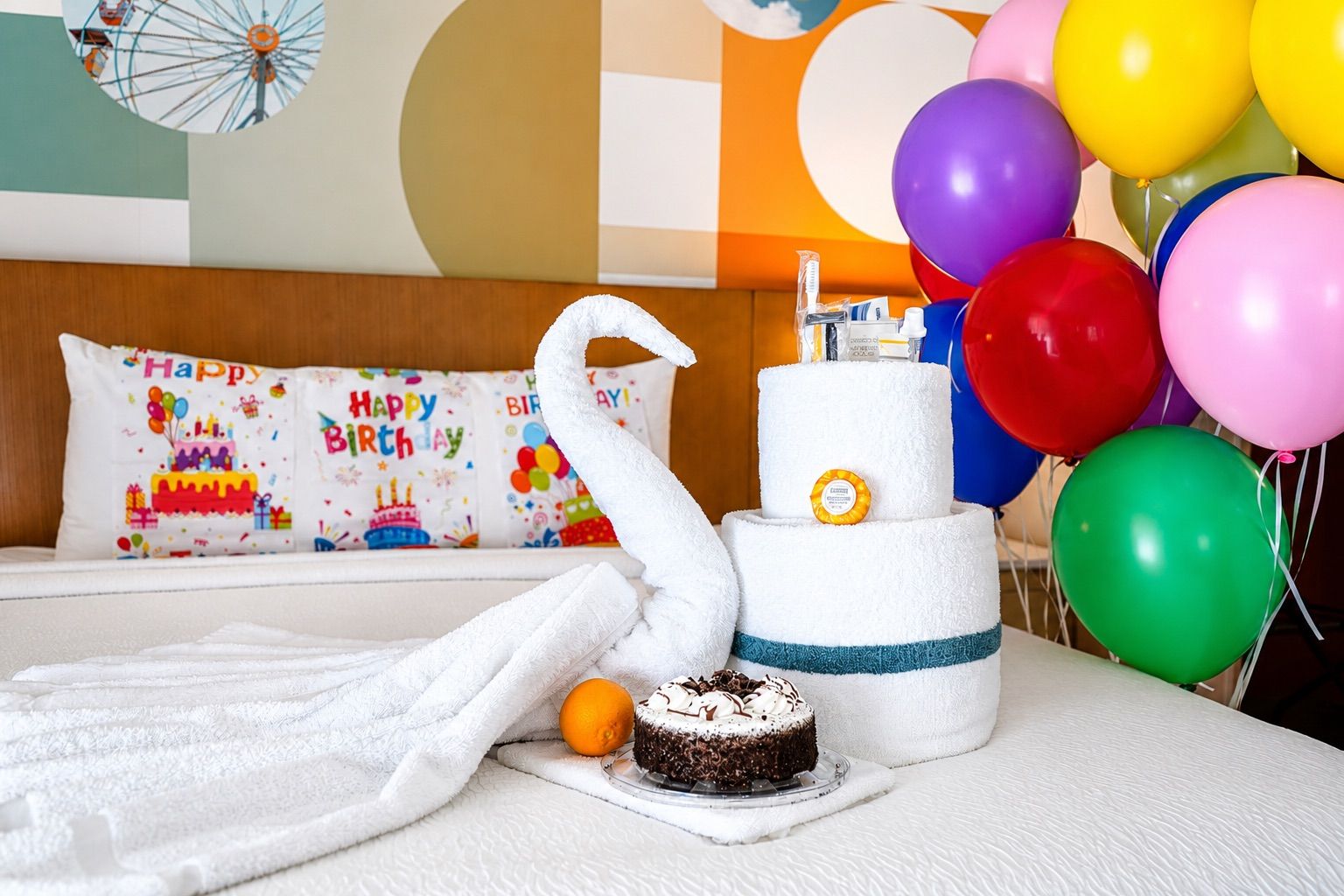A hotel room with a bed, a swan-shaped towel, a birthday cake, balloons, and a pillow with 'Happy Birthday' text.