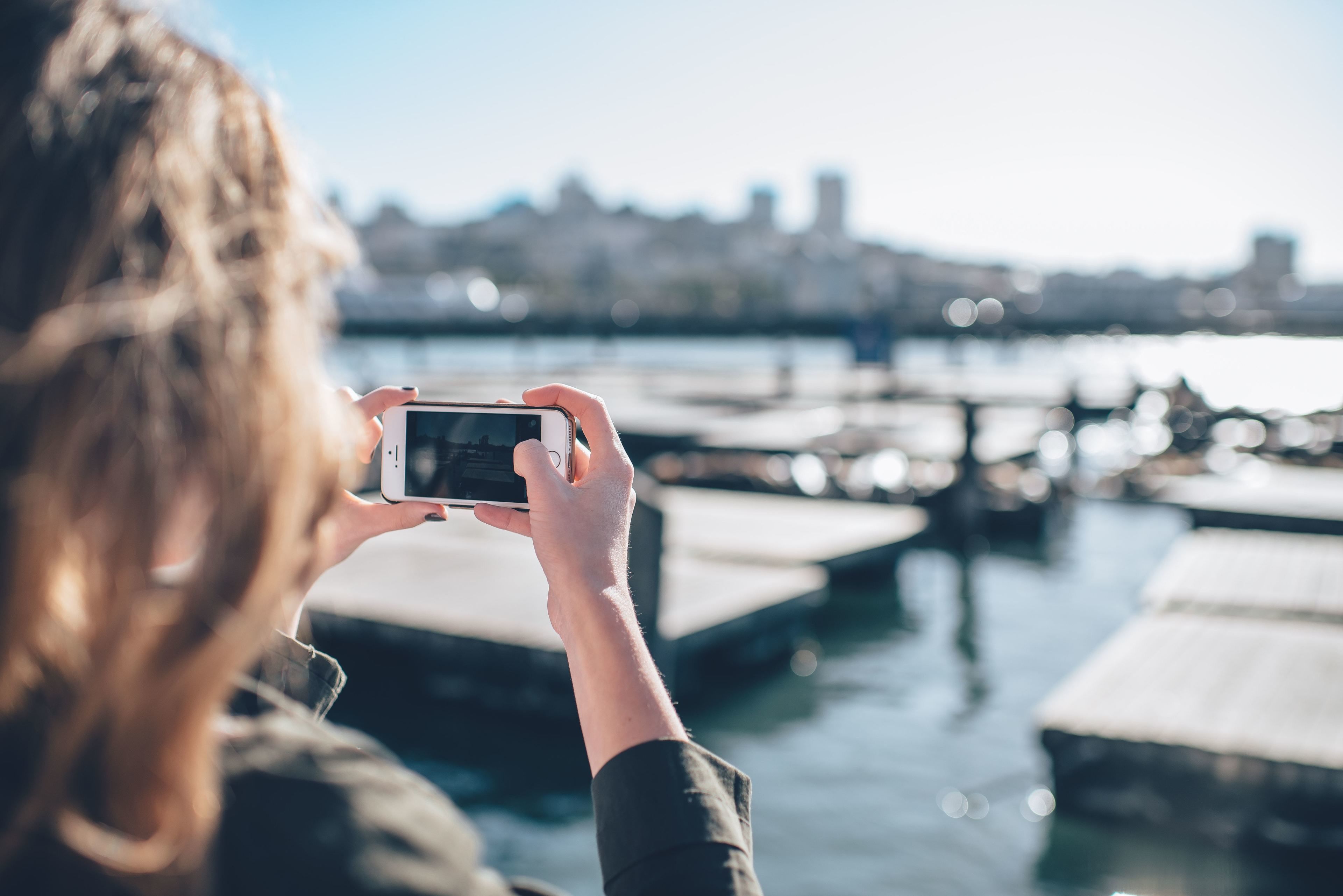A person holding a smartphone, capturing a marina with boats and a city skyline in the background.