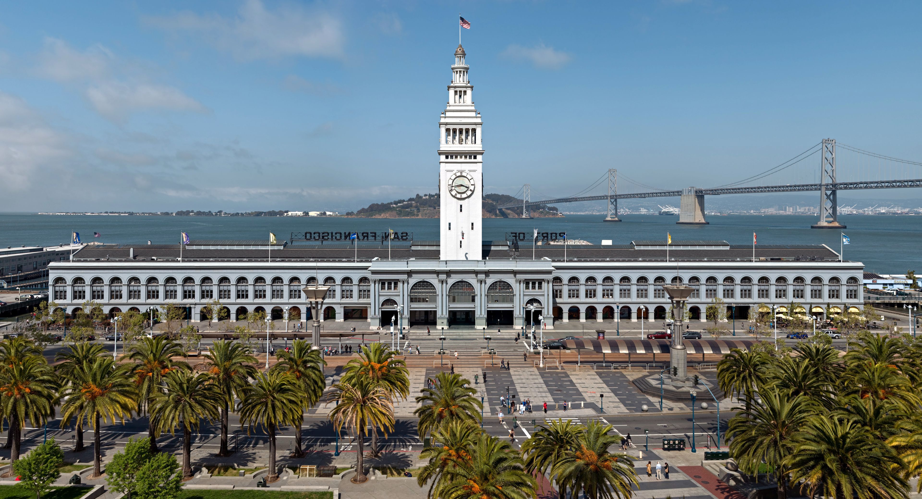 A large white building with a clock tower, palm trees in the foreground, and a bridge in the background.