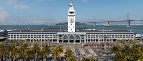 A large white building with a clock tower, palm trees in the foreground, and a bridge in the background.