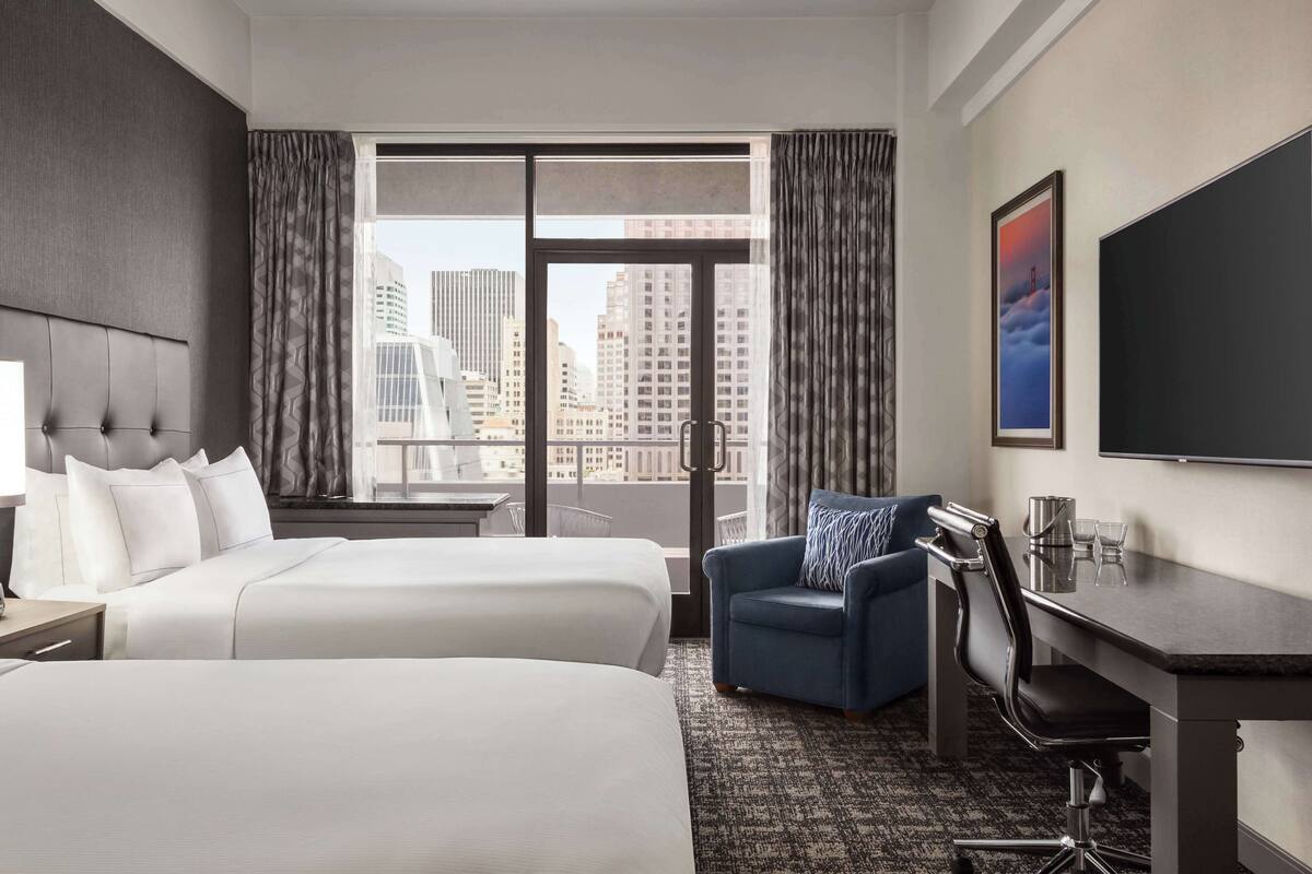 A hotel room with two beds, a desk, a chair, a TV, and a large window with a city view.
