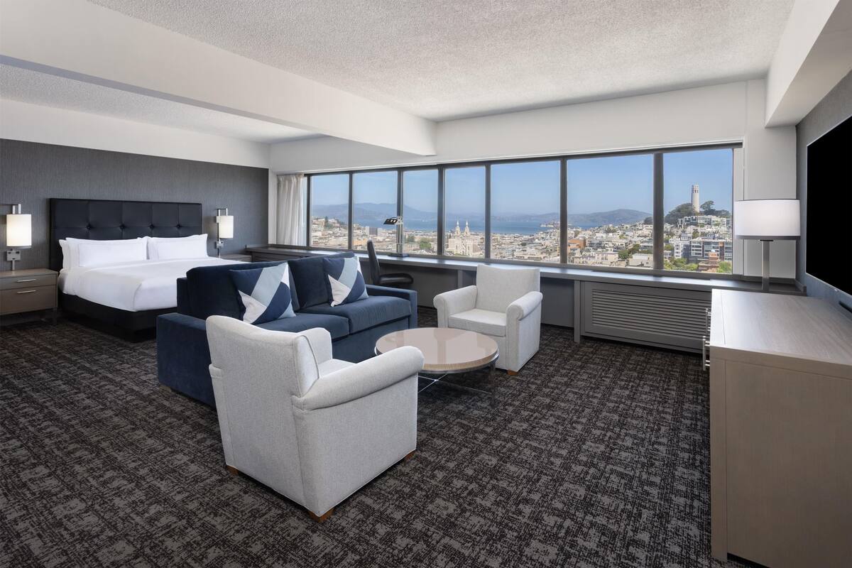 A modern hotel room with a large bed, two sofas, a coffee table, and a TV. The room has a city view through large windows.